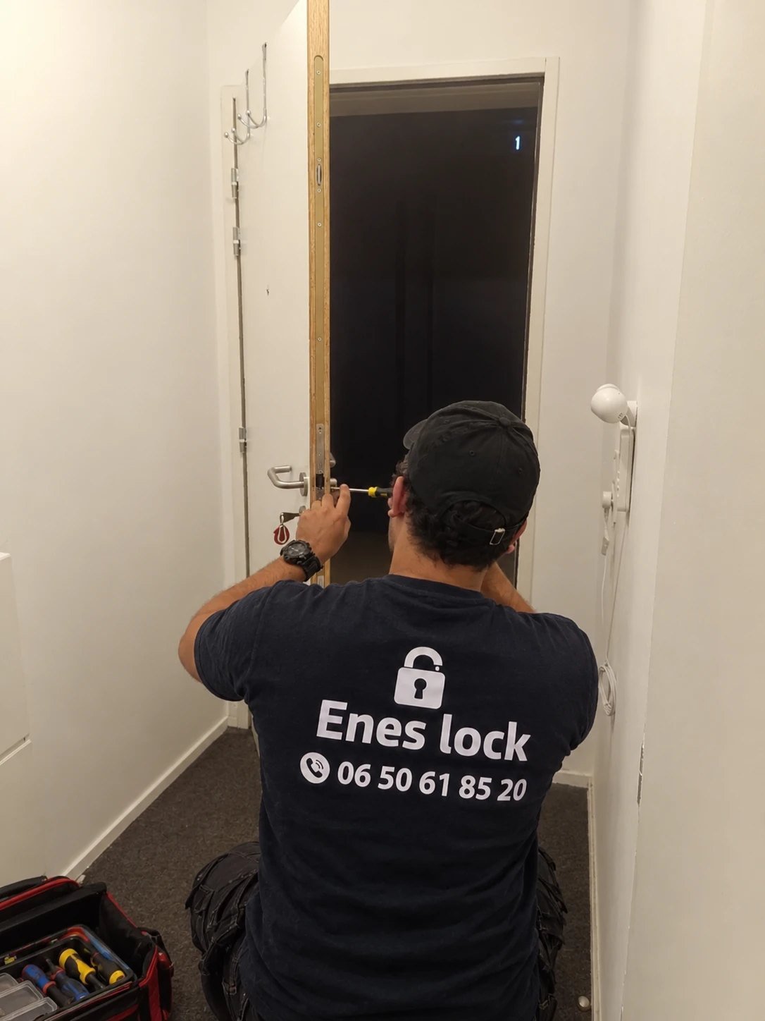 Enes, de dos avec son tee-shirt Enes Lock, règle la serrure 3 points d'une porte d'appartement